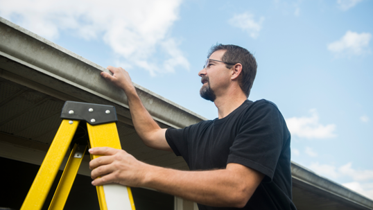 Man on a yellow ladder inspecting the roof gutters